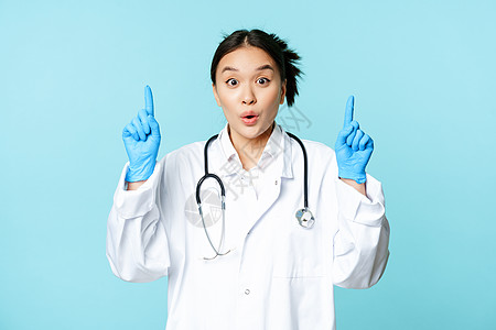 兴奋的亚洲籍医务工作者 女医生举起指尖 佩戴消菌手套和制服 站在蓝背景上站立隔离临床情况女性女士诊所成人专家症状肺炎图片