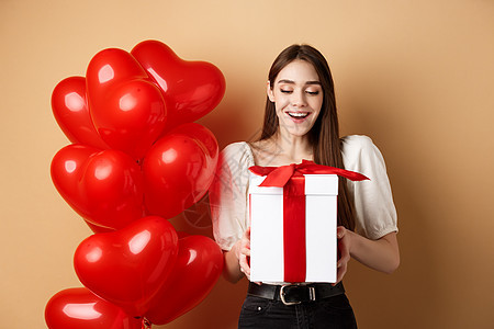 快乐的女士打开情人节礼物 微笑兴奋和看着现在的盒子 站在红心气球旁边 在蜜蜂背景上惊喜浪漫庆典化妆品情人女性女孩伙伴嘴唇男朋友图片
