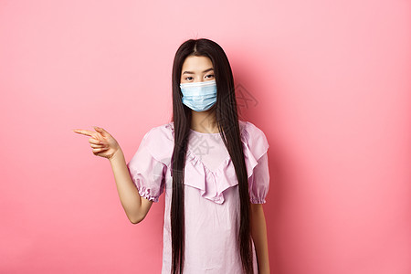 年终促销Covid19 流行病生活方式概念 戴着医用面具的可爱亚洲少女指着左边的标志 展示广告 穿着粉红色背景的裙子情感工作室微笑社交伙背景