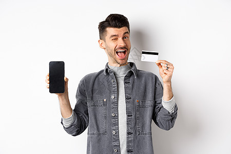 开朗的帅哥展示优惠 空荡荡的智能手机屏幕和塑料信用卡 向你眨眼微笑 推荐报价 白色背景成人技术广告商业男性办公室情绪工作室电子商图片