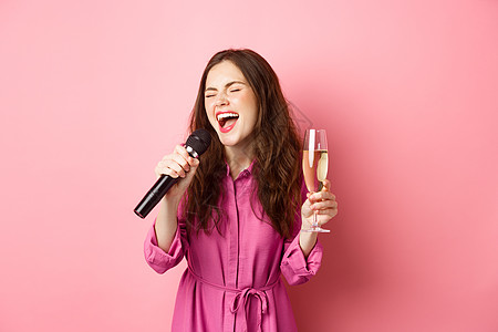 庆祝和节假日概念 派对女性有乐趣 喝香槟 唱卡拉OK 拿着麦克风 在粉红背景下快乐地站立的女士形象工作室广告庆典演讲成人喜悦女朋图片