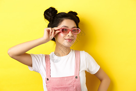 美丽的亚洲女孩穿上时尚的太阳镜和微笑 穿着潮流的夏日服装 站在黄色背景上嘴唇头发女性口红假期情感发型青少年化妆品魅力图片