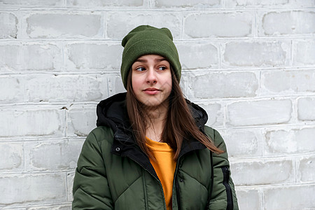一个穿着黄色毛衣和卡其帽的漂亮女孩的肖像 它站在白色砖墙附近 情感的概念 情绪概念图片
