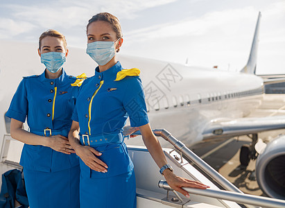 两位身穿蓝色制服 戴着防护面罩的漂亮空中小姐看着镜头 在登机时站在楼梯上图片