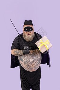 穿着佐罗西装超重的成熟男人惊讶地指着紫色背景的礼盒图片