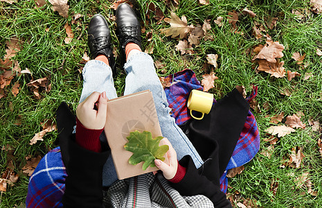 在一个阳光明媚的温暖秋日 在绿色草地上 一个女人拿着一本合上的书躺在她的腿上 上面有一片落下的橡树叶 放松 阅读和放松的概念 顶图片