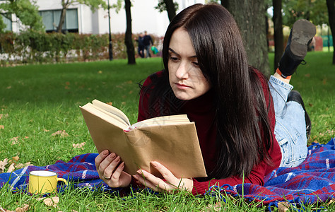 在阳光明媚的日子里 一位年轻女子躺在绿色草地上的城市公园里 躺在羊毛格子毯或毯子上阅读她最喜欢的书 娱乐和休闲 教育和学习的概念图片