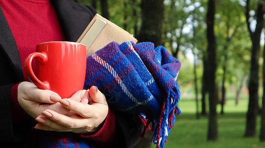 公园里一位穿着毛衣和黑色外套的女人手里拿着一杯红咖啡和一本书 上面有蓝色方格羊毛毯或格子花呢 温暖和晴朗的天气 柔软舒适的摄影橙图片