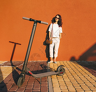 站在墙边的女人 前面有电动摩托车城市女性衣服太阳镜技术黑发女士白色微笑运输图片