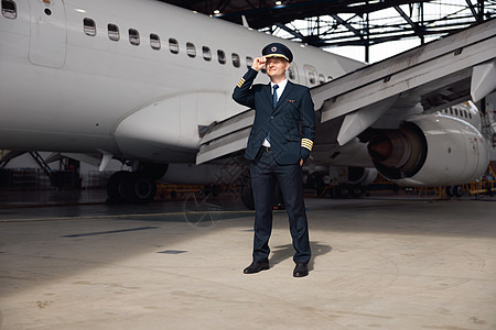 身穿制服 自信的飞行员全长镜头望向远方 调整帽子 站在机场机库的大型客机前图片