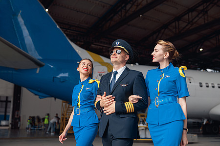 在机场机库的大型客机前 身着制服和飞行员太阳镜的快乐飞行员与两名身着蓝色制服的空姐一起行走精英航班车辆全体旅行飞行航空职业管理人图片