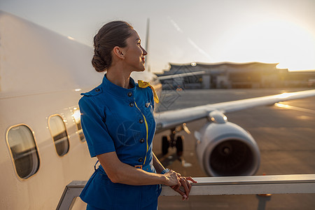 穿着蓝色制服 体贴的空姐站在户外看日落 背景中机场航站楼附近的商用飞机服务女性服务员沉思思维冒充成人楼梯职业微笑图片