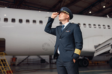 身穿制服的鼓舞人心的飞行员望向远方 调整帽子 站在机场机库的大型客机前图片