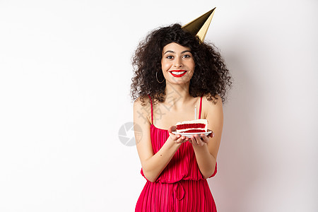穿着红色连衣裙 戴着派对帽庆祝生日 拿着生日蛋糕许愿 对着镜头微笑 站在白色背景上的漂亮女人浪漫假期化妆品裙子投标发型卷曲促销黑图片
