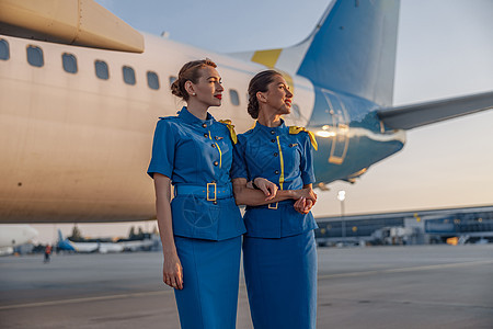 两个穿着蓝色制服的美丽的空姐 笑着微笑着离开 在日落时站在机场一架大型客机前图片