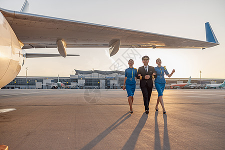 兴奋的男性飞行员和两名身穿蓝制服的女性空服女乘务员在日落时候终点站飞机前行走 一整拍全镜头团队男人空姐职业飞机场人员旅游喷射全体图片