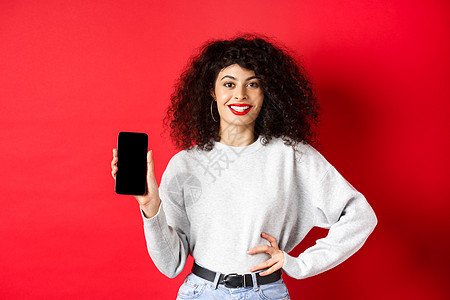 站在红色背景下 微笑着的迷人女性展示着空荡荡的智能手机屏幕 看起来很开心 为在线商店或应用做广告卷曲工作室技术广告购物生活女孩电图片