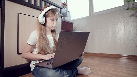 一个聪明的七岁小女孩戴着白色耳机 手里拿着一台笔记本电脑 正在她房间的地板上推来推去 互联网和 IT 技术上的年轻一代图片