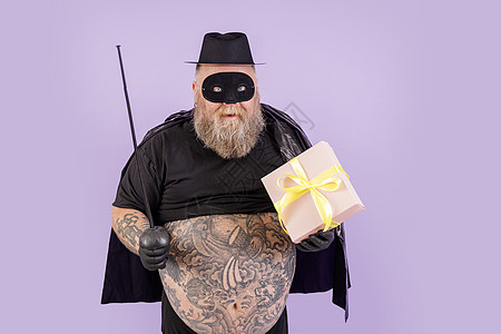 身穿佐罗西装的快乐丰满绅士在紫色背景上拿着玩具重剑和礼盒图片
