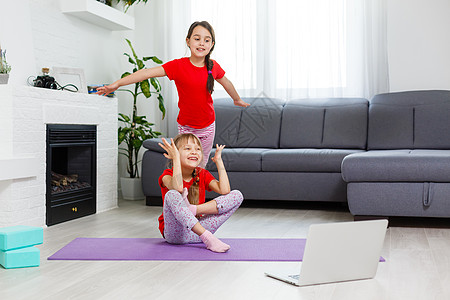 两个小女孩在笔记本上通过视频练习瑜伽 伸展运动和健身 远程在线教育培训 在家有氧 健康的生活方式 冠状病毒 待在家里 孩子们进行图片