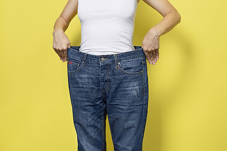 饮食的概念 适当的营养 减肥 展示宽松牛仔裤和她减肥的苗条女人 黄色背景中突显的女式白色 T 恤和超大号牛仔裤腹部女孩活动女性尺图片