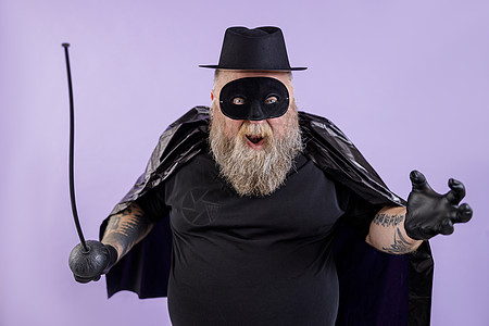 留着胡须的成年男子 体重超重 穿着佐罗套装 紫色背景上摆着玩具重剑图片