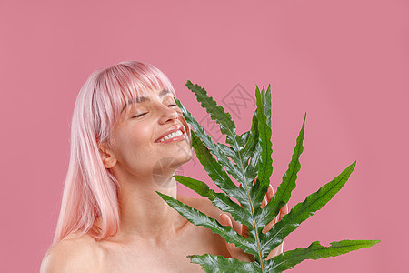 一头粉红色头发 闭着眼睛微笑 手拿植物叶子靠近脸 在粉红色工作室背景中孤立地摆姿势的快乐女人的画像图片