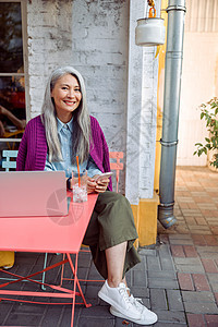 穿着温暖紫色外套的微笑成熟亚洲女士 用笔记本电脑坐在桌子上用手机挂着电话图片