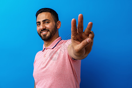 年轻阿拉伯男子笑着 在蓝色背景下举三只手指图片