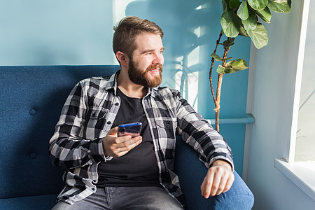 技术与休闲概念在家使用智能手机的帅哥沙发成人胡须快乐男性阅读衬衫长椅检查倾斜图片
