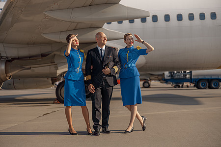在阳光明媚的日子里 快乐飞行员与两名身穿亮蓝色制服的空姐站在飞机前的全长照片图片