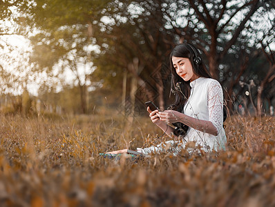 妇女有耳机 听从智能电话的音乐和坐在公园里裙子闲暇森林手机长椅花园女孩日落女性女士图片