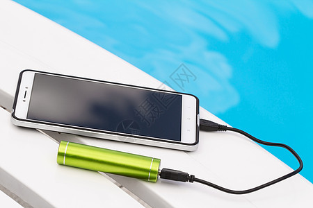 智能手机通过蓝色水背景上的 USB 电缆连接到绿色便携式电池充电器 休息时在游泳池附近的躺椅上为手机紧急充电图片