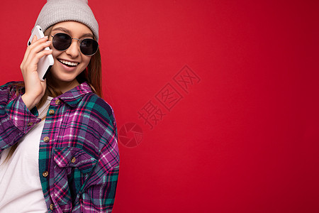 特写照片 美丽快乐 积极向上的年轻金发女性穿着时髦的紫色衬衫和休闲的白色 T 恤灰色帽子和太阳镜 在红色背景中被隔离 手牵着手 图片