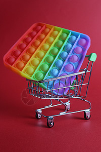 这个多色玩具是一个 popit 简单的凹坑抗压玩具 位于红色背景的一篮婴儿车购物车中 带有文本复制空间 购物手指乐趣艺术塑料流行图片