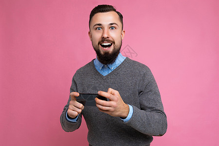 一张快乐英俊的年轻黑发未剃须男子的照片 留着胡子 穿着日常灰色毛衣和蓝色衬衫 隔离在背景墙上 手持智能手机通过手机玩游戏 看着相图片