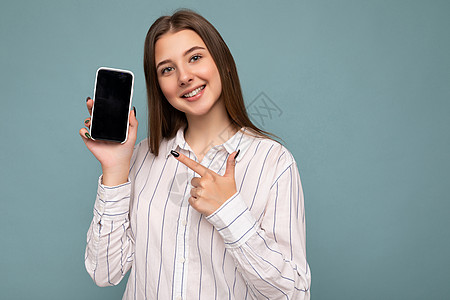 照片中 美丽微笑的年轻女性穿着休闲时尚的服装 看起来很漂亮 站在背景中 与复制空间保持智能手机 手拿手机 屏幕显示为空屏幕 用于图片