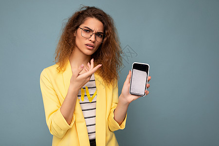 不满 愤怒 迷人的年轻女性 一头卷曲的深色金发 身穿黄色夹克 头戴光学眼镜 在背景中被隔离 手持并展示手机 手机上有空白空间 可图片