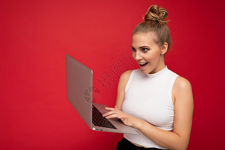 侧脸照片拍摄美丽快乐惊奇惊讶喜出望外的金发年轻女子 头发聚集 穿着白色 T 恤 使用电脑笔记本电脑在键盘上打字 看着红墙背景上孤图片