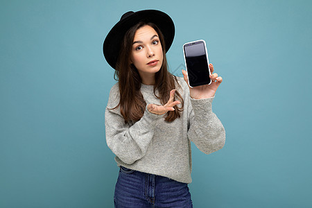 戴着黑帽和灰色毛衣的漂亮年轻女性拿着智能手机 看着背景中突显的相机 提出不满的褐发女性上网电话技术联系屏幕工具细胞游戏互联网青少图片
