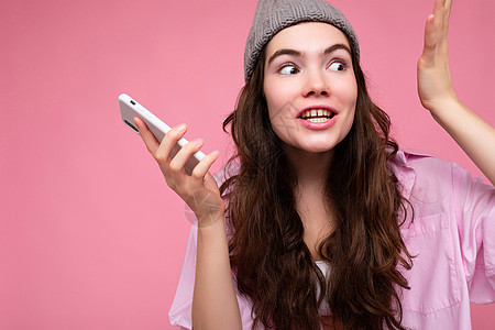 特写照片 情绪迷人的年轻黑发女性穿着时尚的粉红色衬衫和灰色帽子 与粉红色背景隔离 手持并使用手机通信和录制语音信息联系屏幕工具幸图片