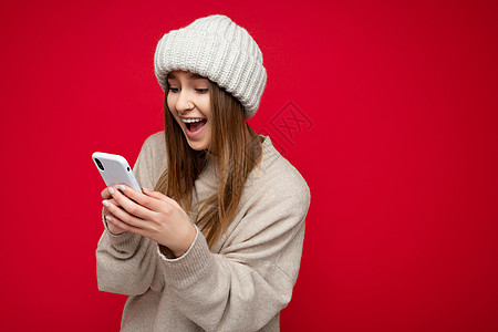 穿着散发美甲毛衣和披风帽的美丽惊人金发年轻女子 因红色背景被手握着 在互联网上使用手机进行在线交流 观看Gadjet展出图片