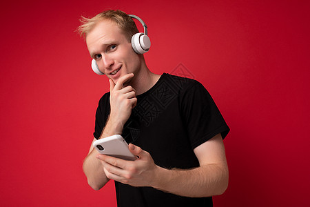 穿着黑色 T 恤和白色耳机的英俊金发年轻人站在红色背景中 拿着智能手机 在网上交流 听着音乐 看着相机玩得开心图片
