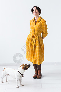 快乐的年轻女子穿着黄色礼服 带着她深爱的狗杰克罗素泰瑞尔站在白色背景上 随意穿戴的概念学生工作室冒充小狗宠物女性猎犬艺术动物友谊图片