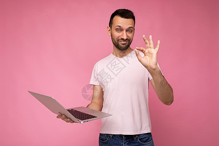 带着笔记本电脑的棕发帅气男子 举着手势 可以看镜头 穿着短袖粉红背景T恤图片