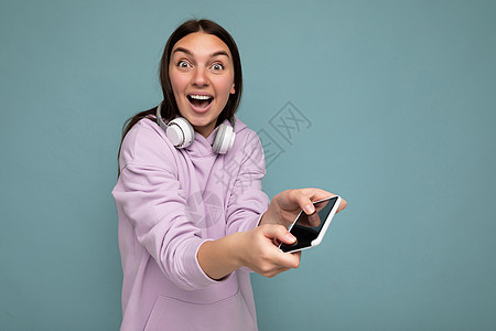 美丽积极的微笑让身穿紫色连帽衫 蓝色背景中突显的深色年轻女性惊讶不已 手持并使用戴着白色无线耳机的智能手机看着相机玩图片