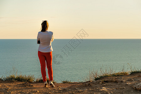 穿着白色夹克和红裤子的年轻漂亮的黑发美女 站在海面悬崖上等待呼唤 后视镜 复制空间 平静 沉默和与自然团结的概念 (笑声)图片