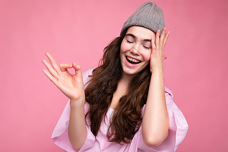 年轻积极快乐美丽的卷发黑发女人的画像 带着真诚的情感穿着时髦的粉红色衬衫和灰色的帽子隔离在粉红色的背景与空旷的空间和显示 ok 图片