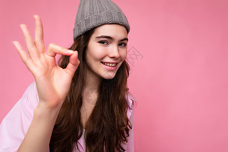 特写成人积极快乐迷人迷人的卷发黑发女人与真诚的情感穿着时尚的粉红色衬衫和灰色帽子隔离在粉红色背景与复制空间并显示 ok 手势 没图片