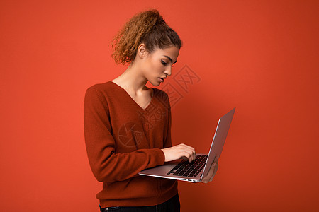 身穿红色毛衣 在红墙背景上隔绝的键盘上的网本显示器打字时 观看着笔记本机的网络监视器背景图片